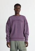 Junger Mann, der einen lila Alpha Industries Pullover und grüne Cargohosen trägt, vor einem schlichten weißen Hintergrund steht.