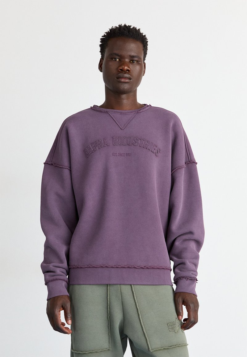 Junger Mann, der einen lila Alpha Industries Pullover und grüne Cargohosen trägt, vor einem schlichten weißen Hintergrund steht.