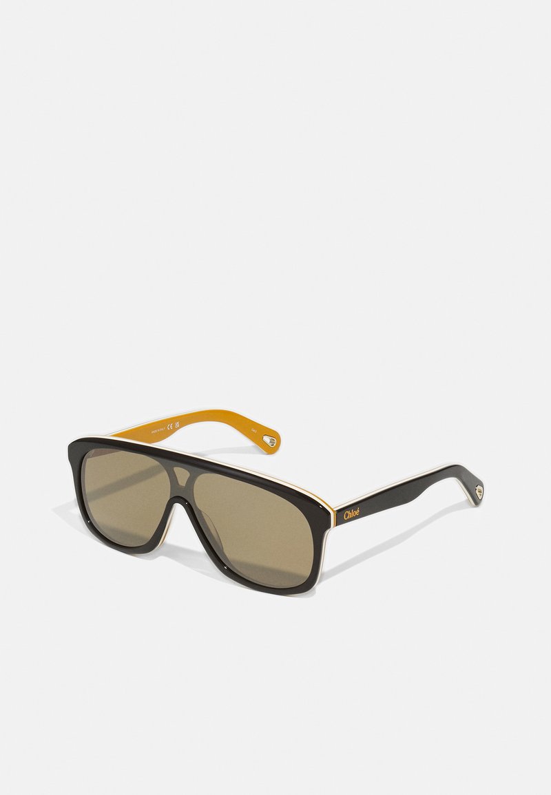 Schwarze übergroße Sonnenbrille mit braunen Gläsern, die an ihrem Rahmen eine weiße Akzentuierung und kontrastierende gelbe klassischen Bügelenden aufweist. Markenlogo vorhanden.