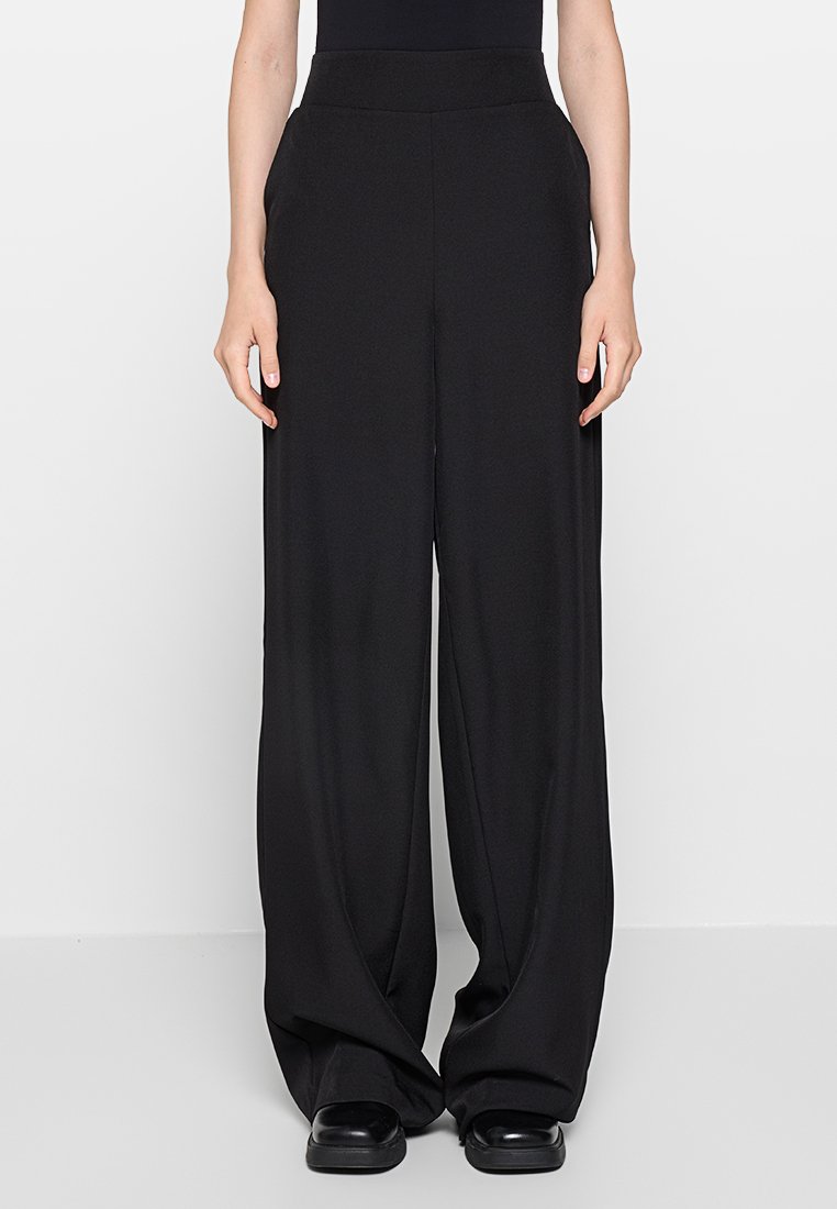 Schwarze, weit geschnittene Hosen mit einer glatten, fließenden Textur. Mit hohem Bund und minimalistischem Design. Getragen mit flachen, schwarzen Schuhen.