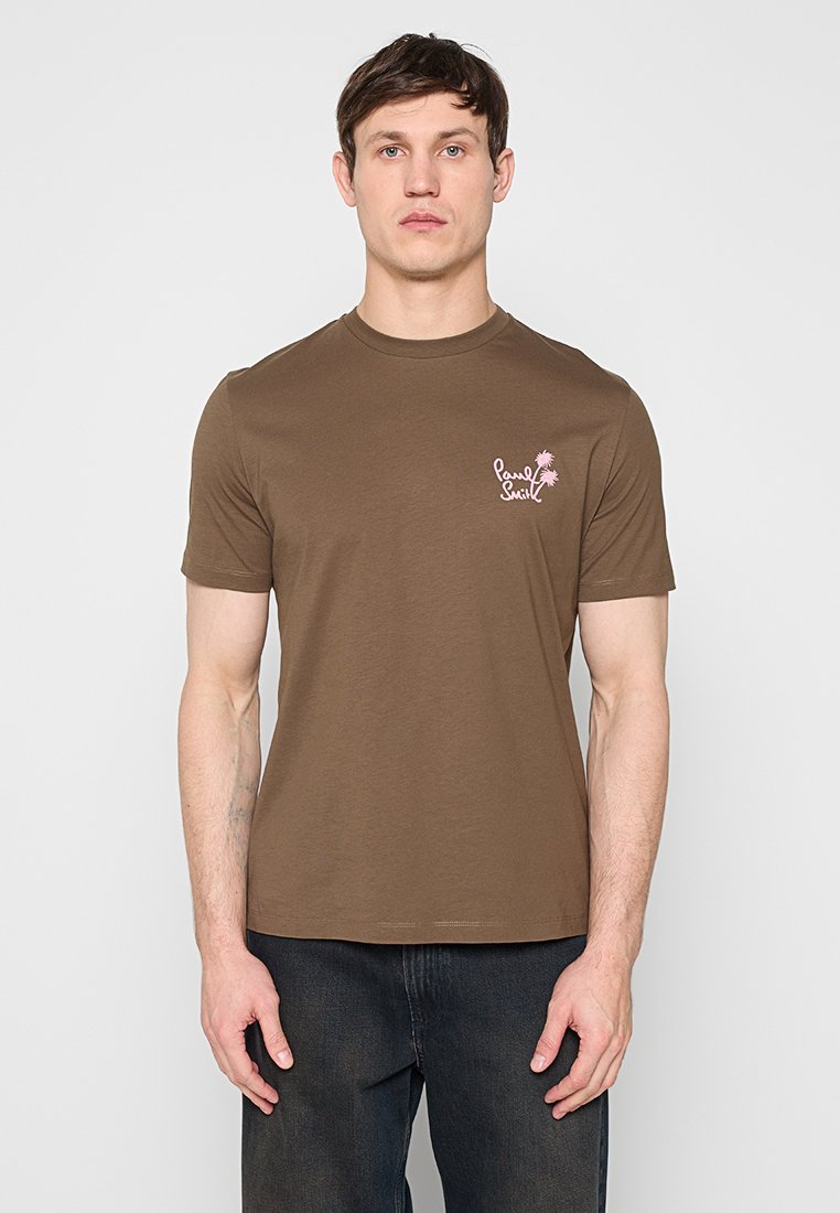 Mann steht und trägt ein braunes T-Shirt mit kurzen Ärmeln, auf dem in Pink "Paul Smith" neben einer Palmen-Grafik in Brustnähe zu sehen ist, kombiniert mit dunklen Jeans.