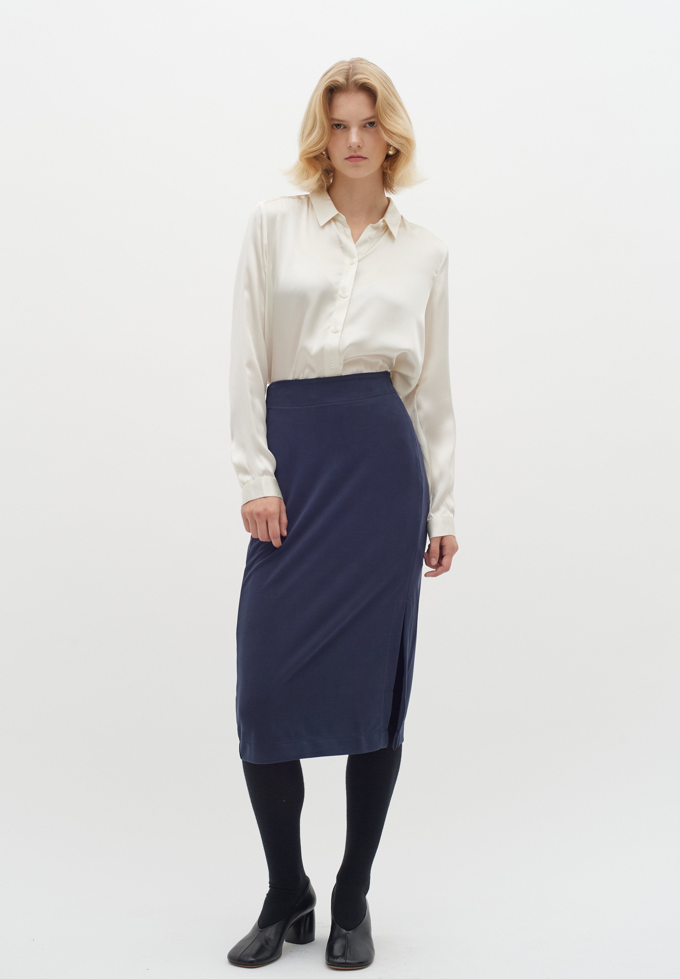 Cremfarbene Satinbluse mit Knopfleiste, kombiniert mit einem marineblauen Bleistiftrock. Schwarze Strumpfhosen und Absatzschuhe runden das Outfit ab.