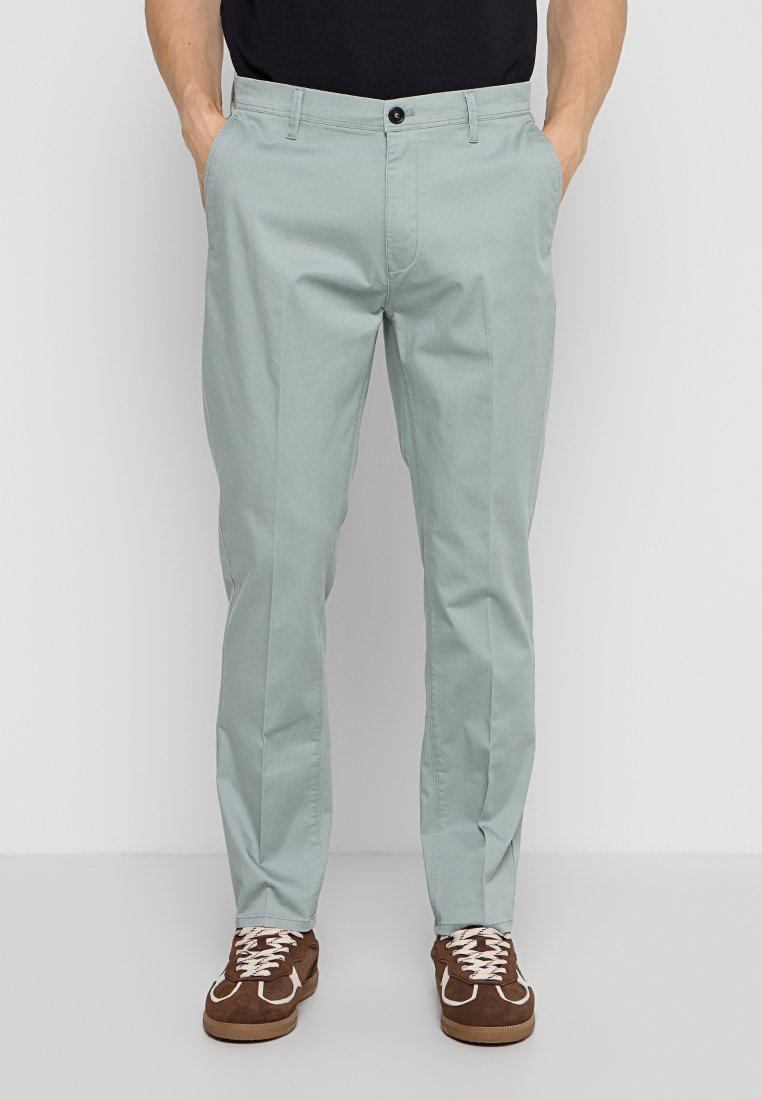Mann trägt hellgraue Slim-Fit-Hose, schwarzes Hemd und braune Sneakers, steht mit Händen in den Taschen.