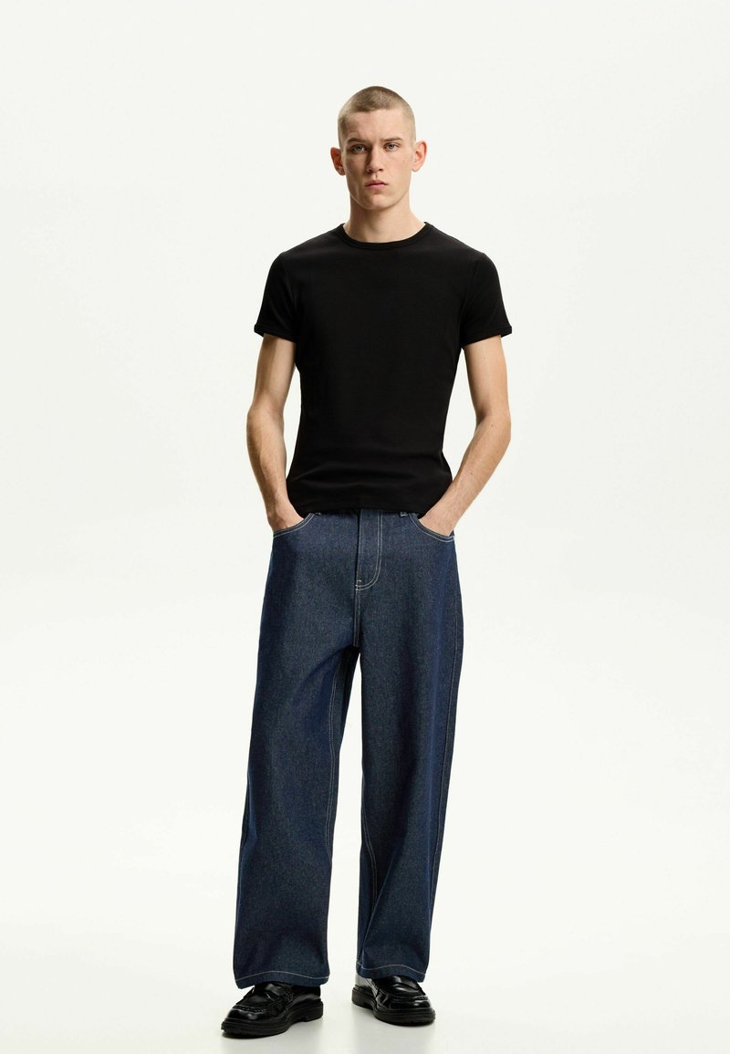 Schwarzes T-Shirt mit kurzen Ärmeln, kombiniert mit weitlegigen Denim-Jeans. Die Jeans sind dunkelblau mit sichtbaren Nähten und einer lockeren Passform. Schwarze Schuhe runden das Outfit ab.