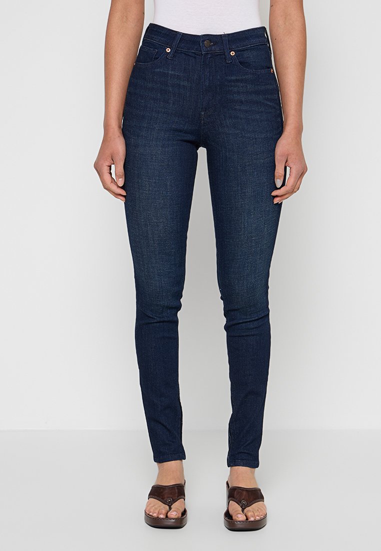 Frau trägt dunkelblaue Skinny-Jeans, steht mit entspannt an den Seiten hängenden Händen, kombiniert mit braunen Sandalen mit offenen Zehen.