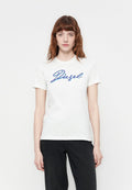 Junge Frau mit roten Haaren, die ein weißes T-Shirt mit Diesel-Logo und schwarze Hosen trägt, steht vor einem schlichten weißen Hintergrund.