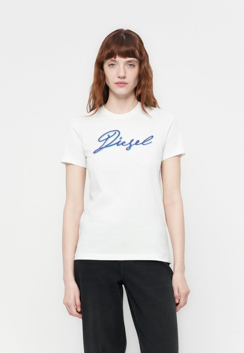Junge Frau mit roten Haaren, die ein weißes T-Shirt mit Diesel-Logo und schwarze Hosen trägt, steht vor einem schlichten weißen Hintergrund.