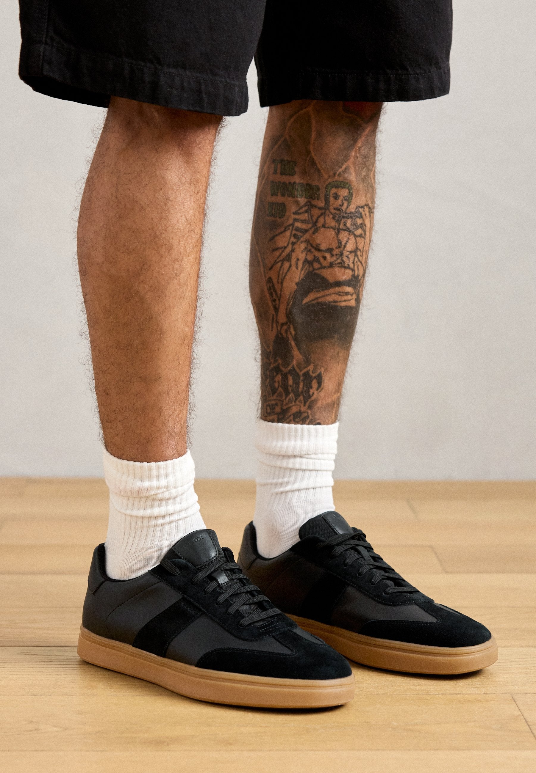 Schwarze Wildleder- und Syntheseschuhe mit einer Gummisohle, die mit strukturierten Einsätzen und Schnürsenkeln ausgestattet sind. Weiße gerippte Socken, die über dem Knöchel getragen werden.