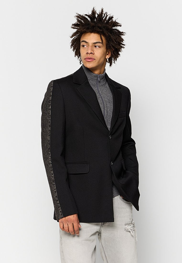 Schwarzer Blazer mit einer strukturierten grauen Wollärmel. Mit einem Reverskragen und zwei Fronttaschen. Getragen über einem grauen Rollkragenpullover.