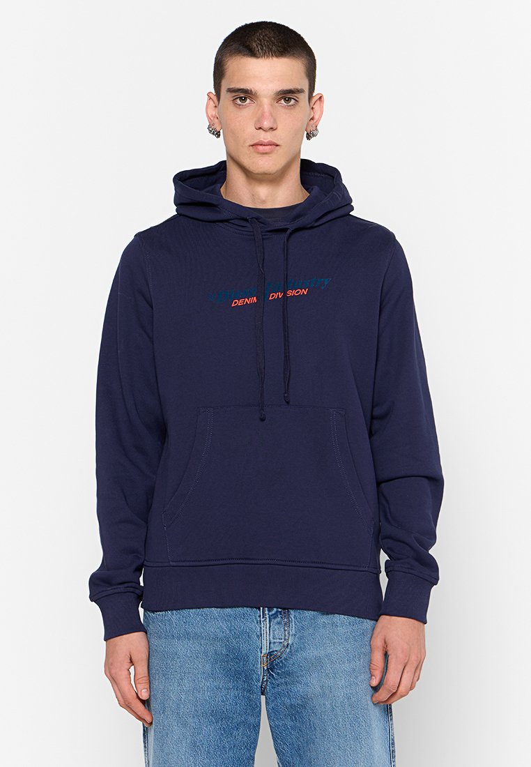 Junger Mann in einem navyblauen Kapuzenpulli mit Fronttasche und blauen Jeans, der vor einem schlichten weißen Hintergrund steht.
