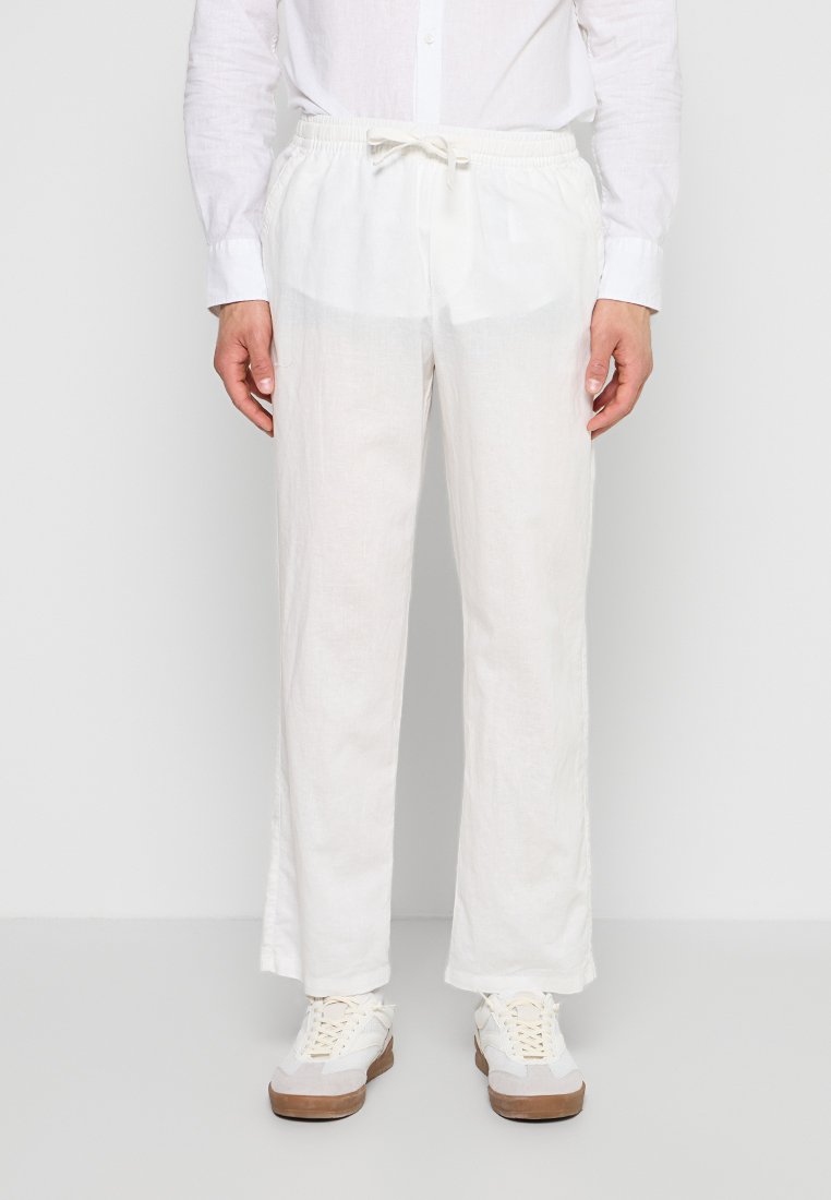 Person trägt lockere weiße Hose mit Kordelzug, weißes Langarmshirt und weiße Sneakers mit Gummisohlen, steht vor einem schlichten Hintergrund.