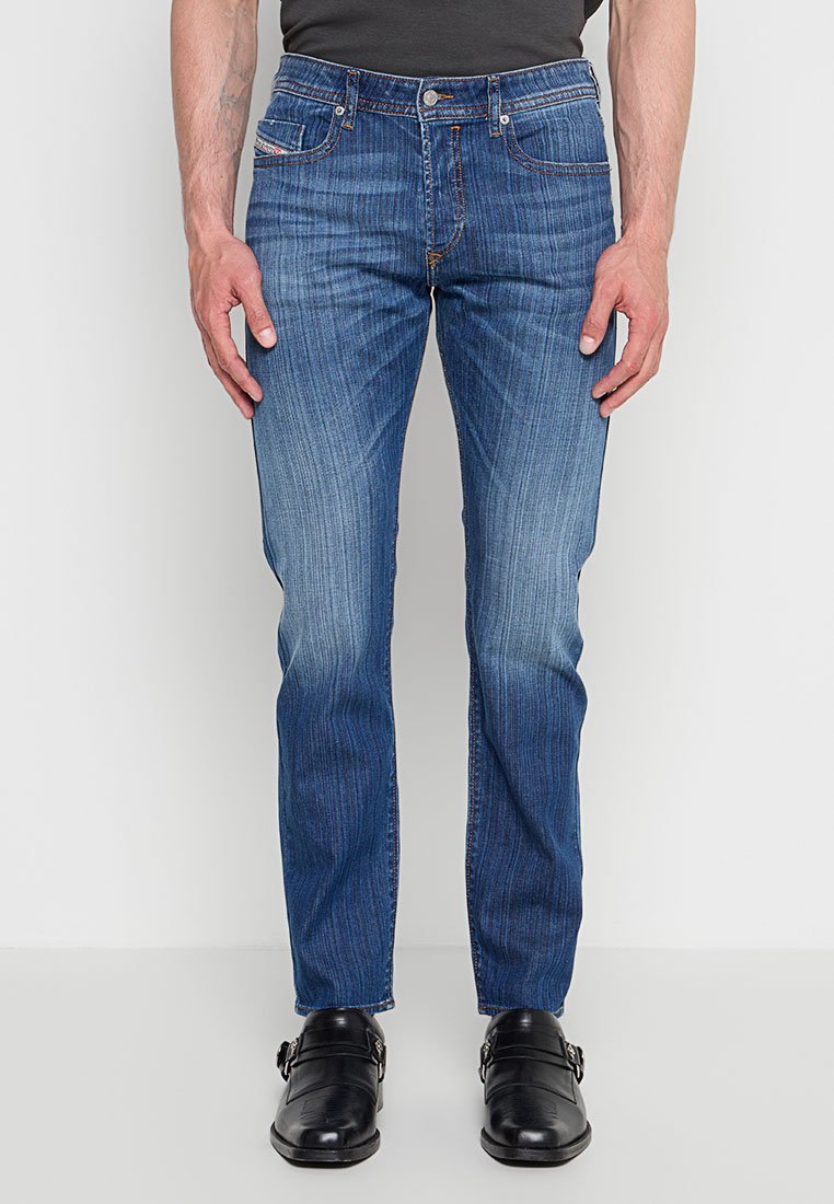 Herren Slim-Fit blaue Jeans mit dezentem Verblassen, getragen mit schwarzen Lederschuhen mit doppeltem Monk-Riemen, stehend vor einem schlichten hellen Hintergrund.
