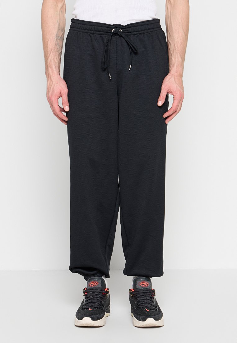 Mann trägt lockere schwarze Jogginghose mit Kordelzug und schwarze Sneakers mit roten Details, steht vor weißem Hintergrund.