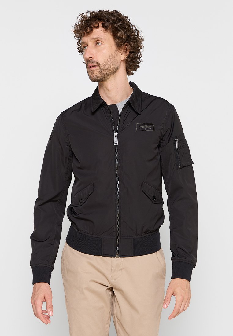 Schwarze Bomberjacke aus leichten Materialien mit einem Reißverschluss auf der Vorderseite, gerippten Bündchen und Saum, ausgestattet mit seitlichen Taschen und einer Tasche am Ärmel.