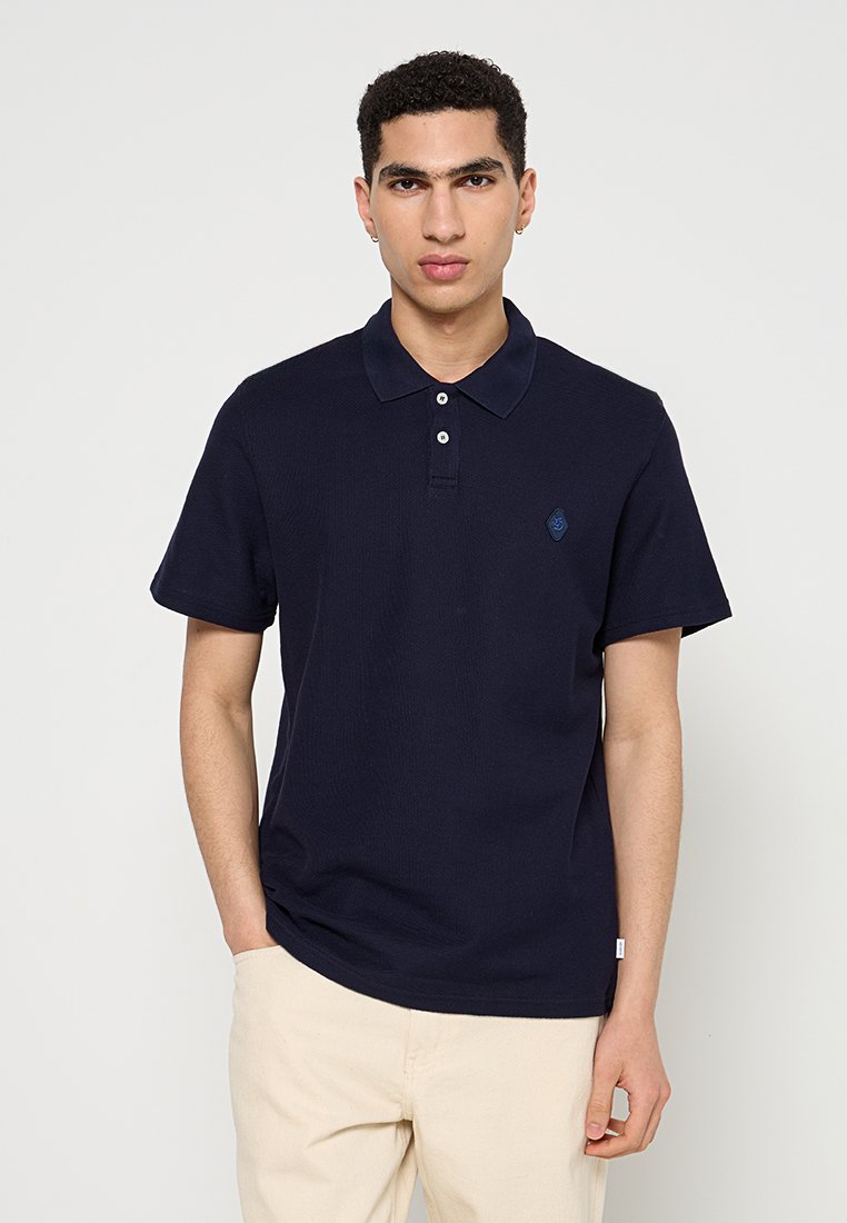 Junger Mann mit kurzen lockigen Haaren, der ein dunkelblaues Polohemd und beige Hosen trägt, steht vor einem schlichten weißen Hintergrund.