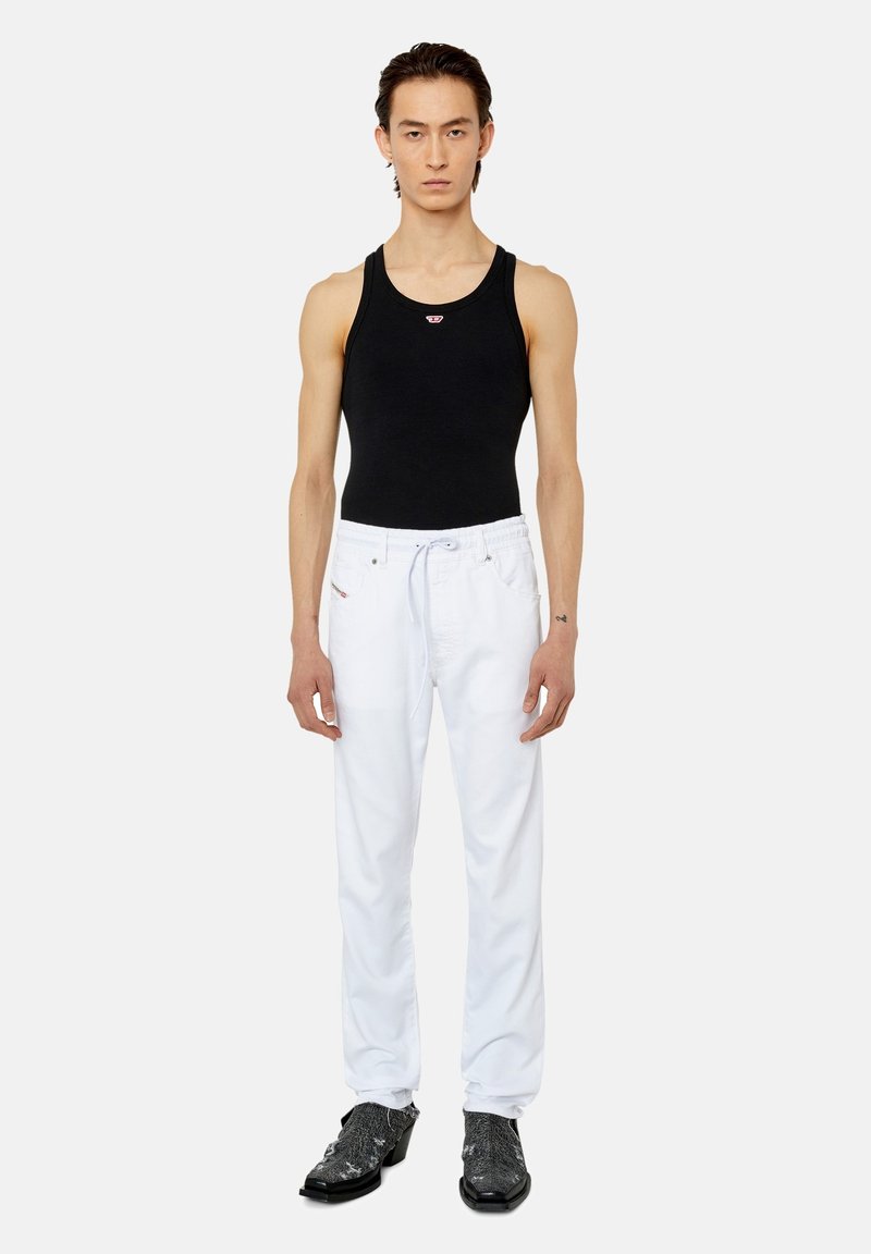 Junger Mann in einem schwarzen ärmellosen Top, weißen Jogginghosen mit Kordelzug und glänzenden schwarzen Stiefeletten, der vor einem schlichten weißen Hintergrund steht.