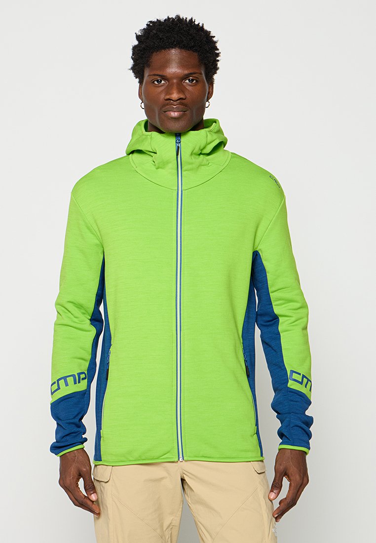 Mann, der eine leuchtend grüne und blaue Zip-up Kapuzenjacke und beige Hose trägt, steht vor einem schlichten hellen Hintergrund.