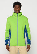 Mann, der eine leuchtend grüne und blaue Zip-up Kapuzenjacke und beige Hose trägt, steht vor einem schlichten hellen Hintergrund.