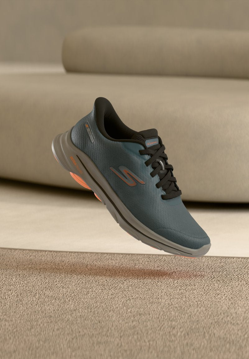 Dunkelgrauer Skechers-Turnschuh mit schwarzen Schnürsenkeln und orangefarbenen Akzenten, der über einem beigen Teppich in einem neutral gehaltenen Raum schwebt.