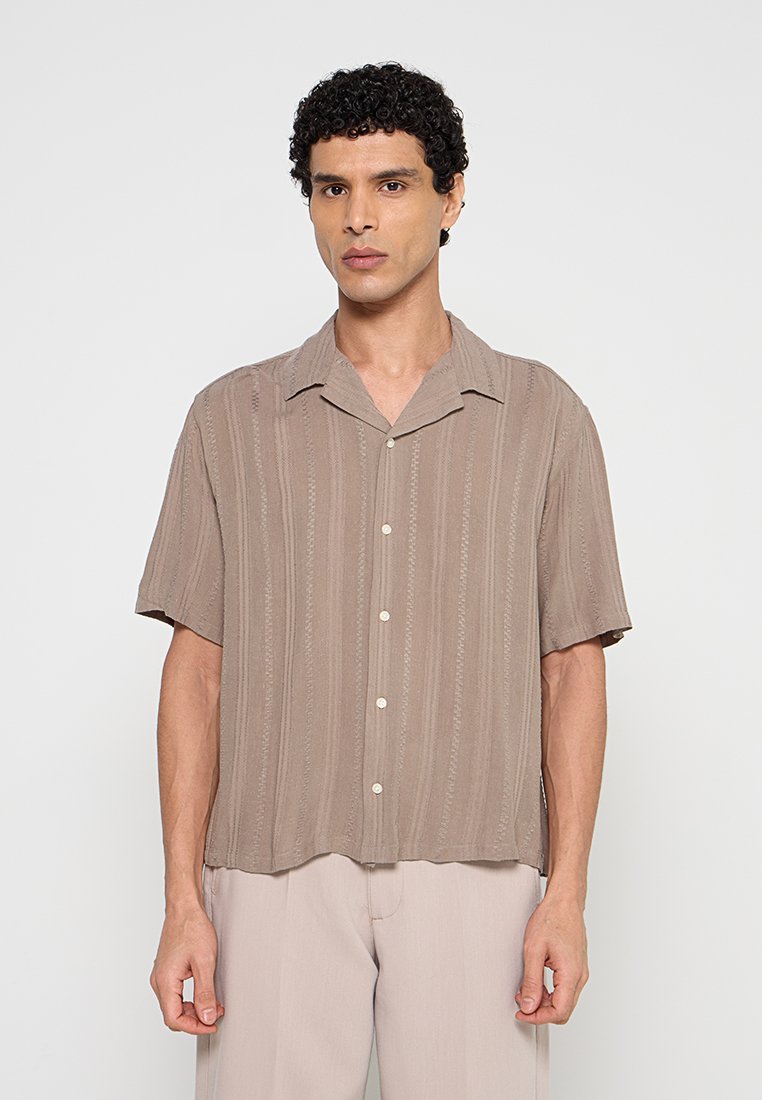 Mann mit kurzen lockigen Haaren, der ein taupefarbenes kurzärmeliges Hemd mit Knöpfen und hellbeige Hosen trägt, steht vor einem schlichten Hintergrund.