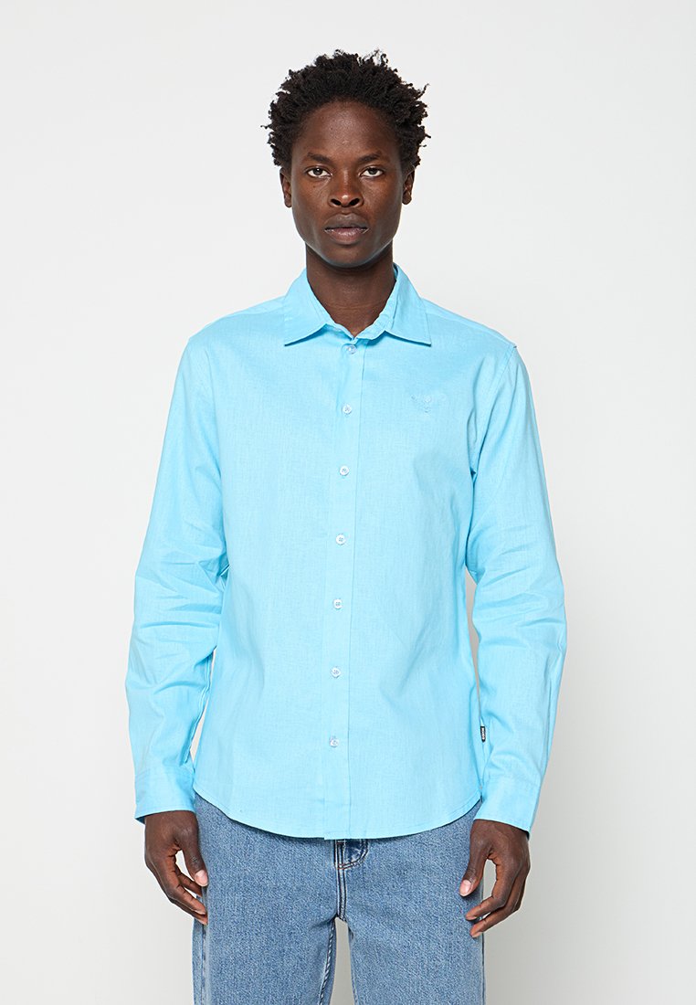 Mann mit kurzen lockigen Haaren, der ein hellblaues langärmliges Hemd und blaue Jeans trägt, steht vor einem schlichten Hintergrund.