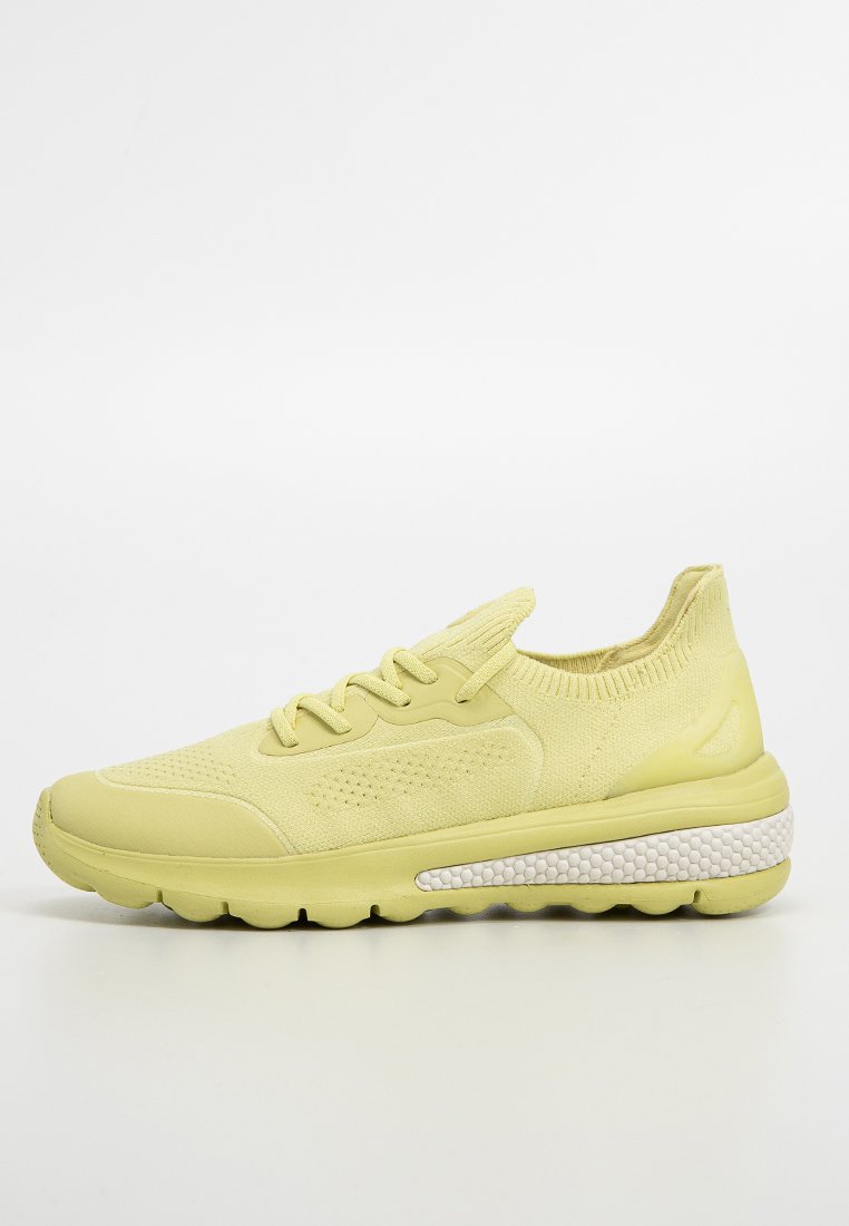 Hellgelbes Sportschuh mit strukturiertem Obermaterial, runder Zehenpartie und einer klobigen Sohle mit einem weißen, gepunkteten Akzentband.