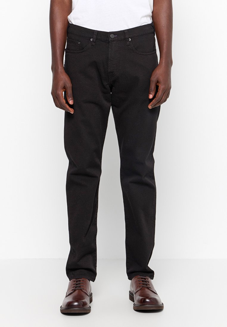 Schwarze Jeanshose mit geradem Schnitt, fünf Taschen und Knopfverschluss. Kombiniert mit braunen Lederschuhen, die eine glatte Textur aufweisen.