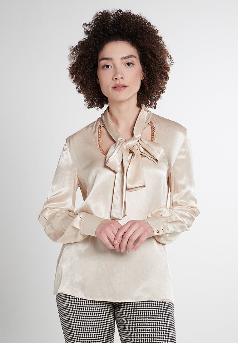 Beige Satinbluse mit einer gebundenen Schleife am Ausschnitt, langen Ärmeln mit Knopfdetails und einem dezenten Glanz. Hahnentritt-Musterhose.