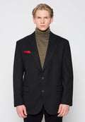 Schwarzer Blazer mit Reverskragen und zwei Knöpfen, kombiniert mit einem gerippten olivgrünen Rollkragenpullover. Mit rotem Akzent auf der Tasche.