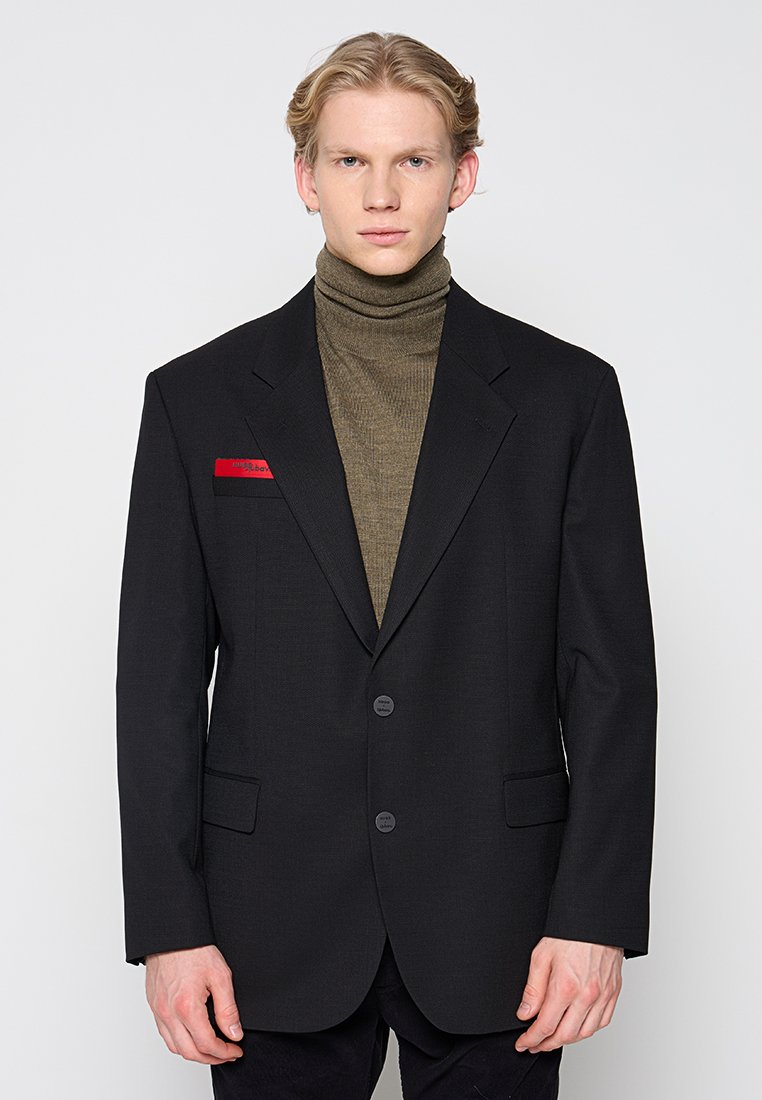 Schwarzer Blazer mit Reverskragen und zwei Knöpfen, kombiniert mit einem gerippten olivgrünen Rollkragenpullover. Mit rotem Akzent auf der Tasche.