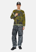 Grüner, gemusterter Strickpullover mit grauen Akzenten, kombiniert mit grauen, stark abgewetzten Cargo-Hosen mit Taschen. Eine schwarze Mütze und Sneakers runden den Look ab.