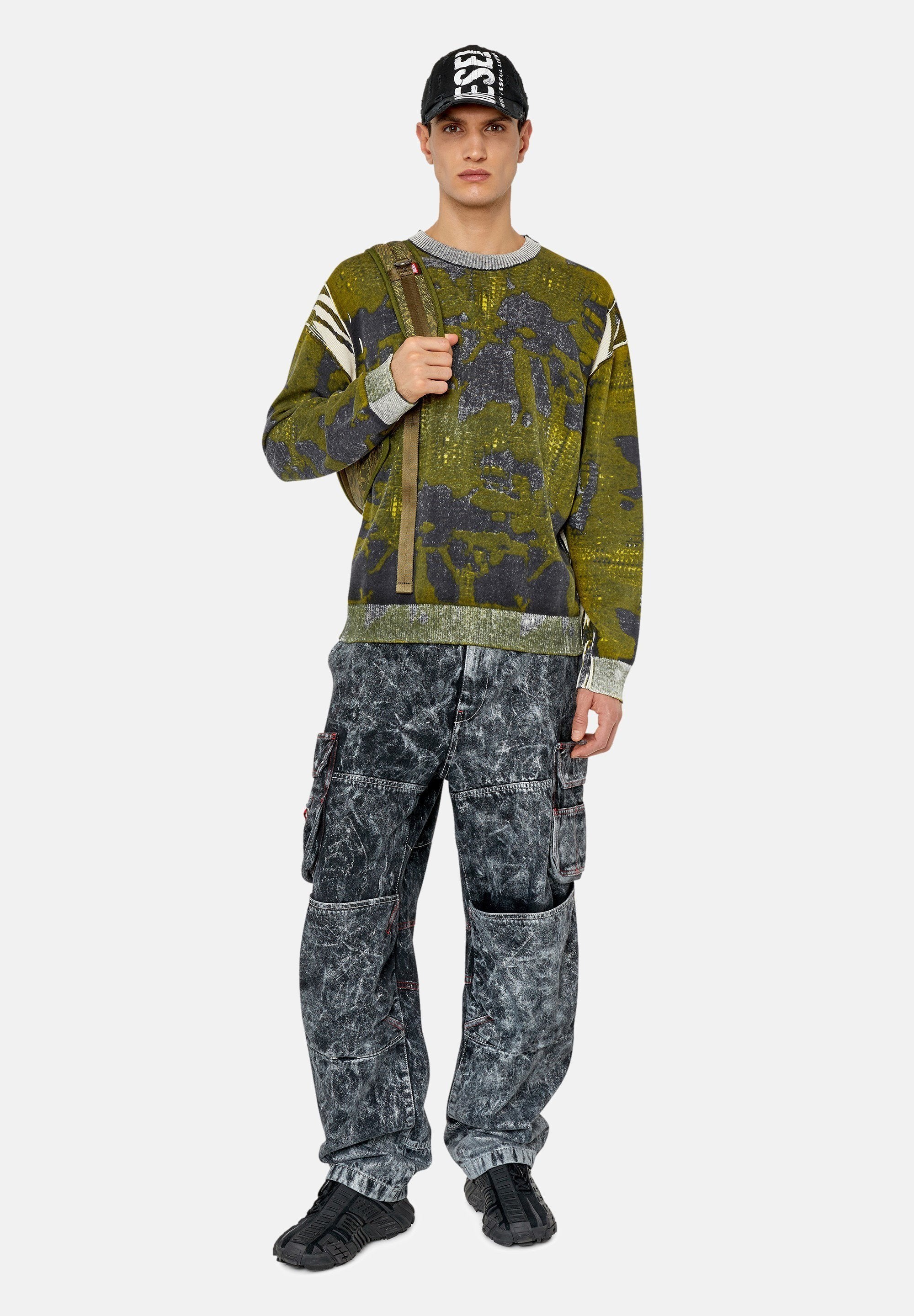 Grüner, gemusterter Strickpullover mit grauen Akzenten, kombiniert mit grauen, stark abgewetzten Cargo-Hosen mit Taschen. Eine schwarze Mütze und Sneakers runden den Look ab.