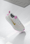Hellgraue Sportschuh mit pinken Akzenten und einem goldenen Swoosh-Logo. Verfügt über ein Obermaterial aus Mesh, eine gepolsterte Zunge und eine strukturierte Sohle.