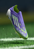 Lila und grüne Fußballschuhe mit strukturiertem Obermaterial, gemustertem Design und multidirektionalen Stollen, über Gras schwebend mit aufgewirbelter Erde.