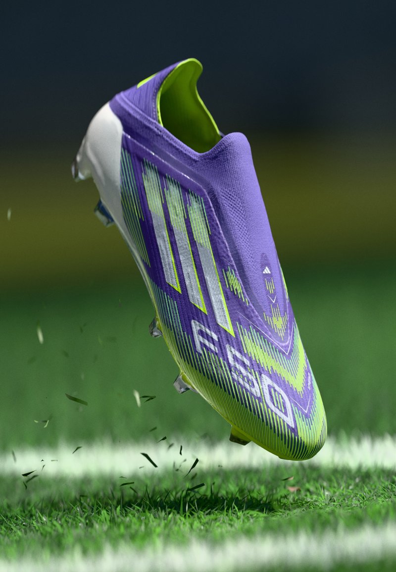Lila und grüne Fußballschuhe mit strukturiertem Obermaterial, gemustertem Design und multidirektionalen Stollen, über Gras schwebend mit aufgewirbelter Erde.