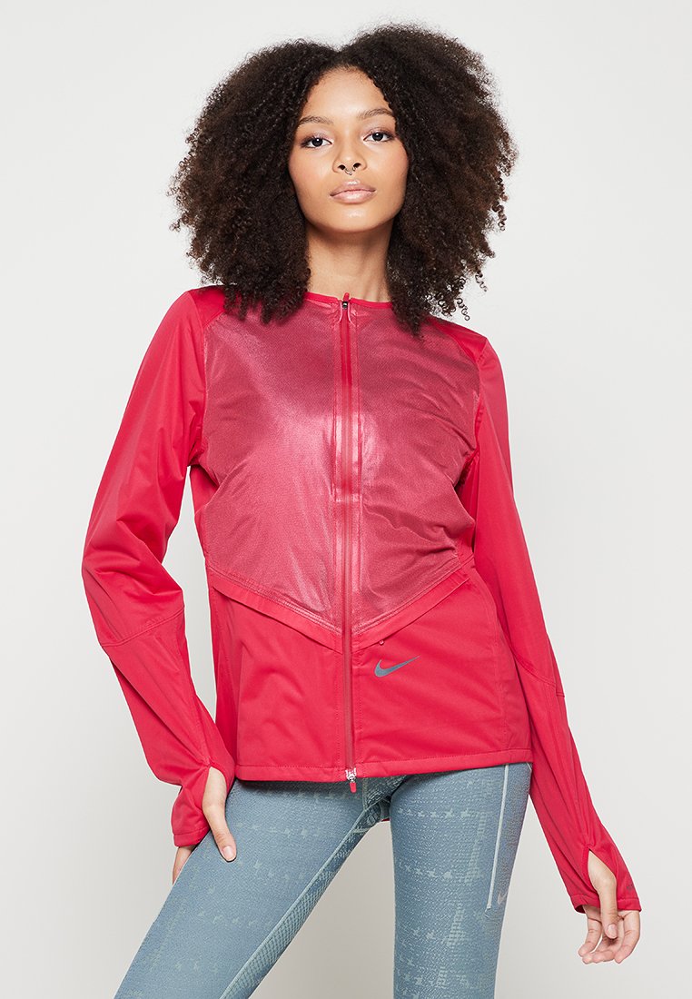 Rote Sportjacke mit Reißverschluss vorne, Netzpaneelen und langen Ärmeln. Mit Nike-Logo und tailliertem Design. Kombiniert mit blauen gemusterten Leggings.