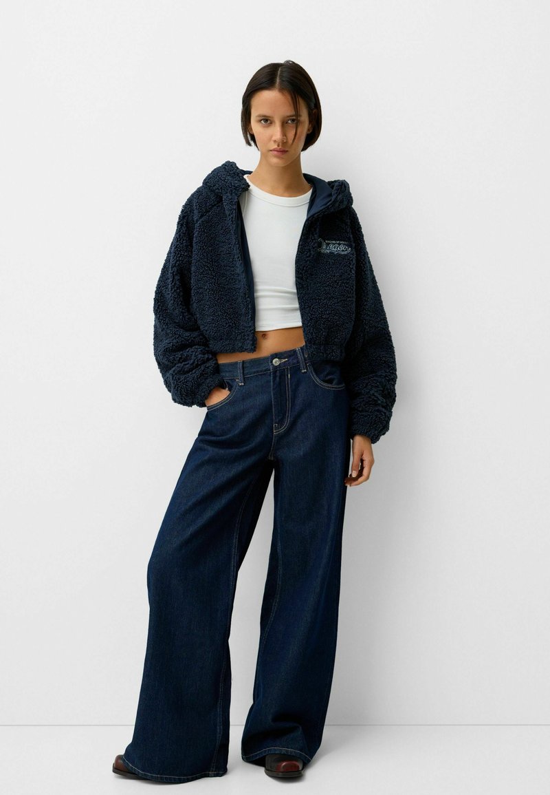 Dunkelblaue, kürzere Fleecejacke mit Kapuze, die ein Logo zeigt, kombiniert mit weit geschnittenen, dunklen Jeans. Beide Kleidungsstücke haben minimale Details.