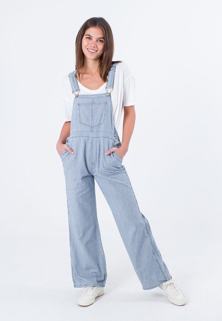 Blaue gestreifte Denim-Latzhose mit weitem Bein, Fronttaschen, verstellbaren Trägern, kombiniert mit einem weißen Hemd und weißen Sneakers.