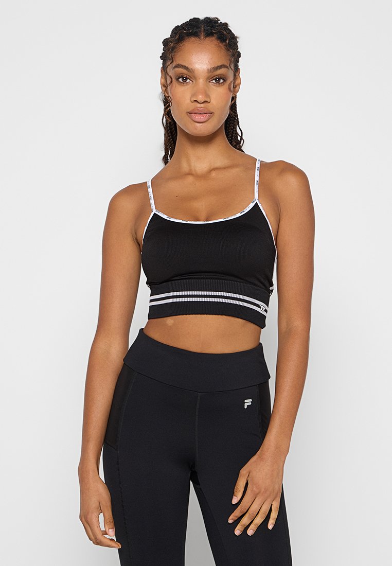 Junge Frau mit geflochtenem Haar, die ein schwarzes Crop-Top mit dünnen weißen Trägern und hohe schwarze Leggings mit Logo auf dem linken Oberschenkel trägt.
