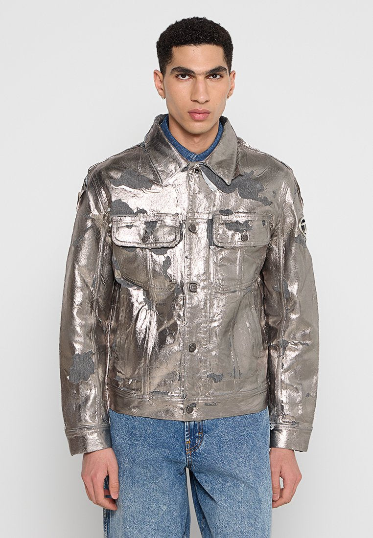 Metallisch silberne Denimjacke mit zwei Brusttaschen, einer durchgehenden Knopfleiste und einer strukturierten Oberfläche. Getragen mit einem blauen Hemd darunter.