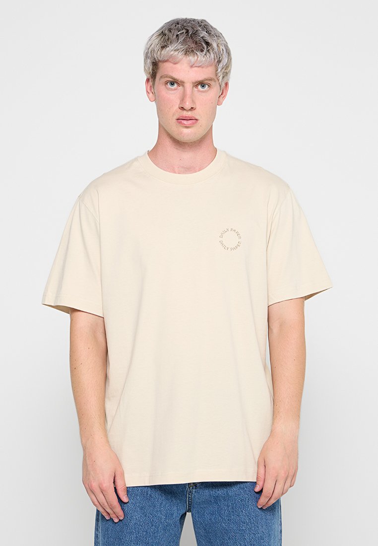 Beige Baumwoll-T-Shirt mit Rundhals und kurzen Ärmeln. Verfügt über ein kleines kreisförmiges Logo, das auf der linken Seite aufgedruckt ist. Getragen mit blauen Denim-Jeans.