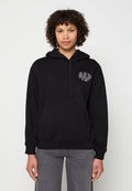 Person mit lockigem Haar, die einen schwarzen GAP-Hoodie und graue Jeans trägt, vor einem schlichten hellen Hintergrund steht.