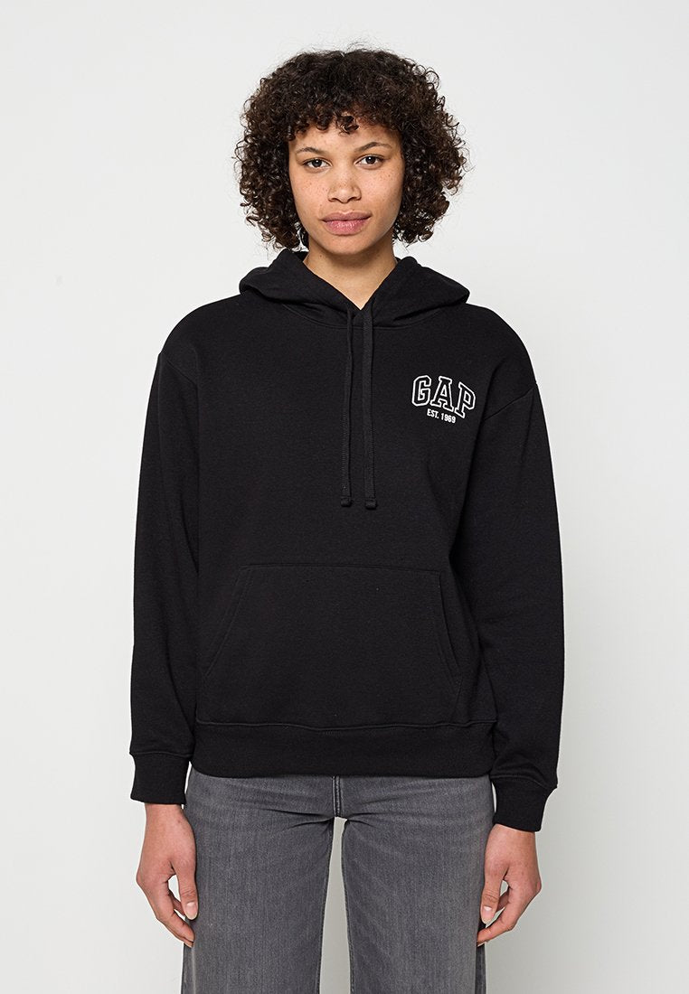 Person mit lockigem Haar, die einen schwarzen GAP-Hoodie und graue Jeans trägt, vor einem schlichten hellen Hintergrund steht.