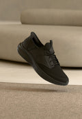 Schwarzer sportlicher Sneaker mit Mesh-Obermaterial und Schnürsenkeln, der über einem beigen Teppich vor einem beigen Sofa schwebt.