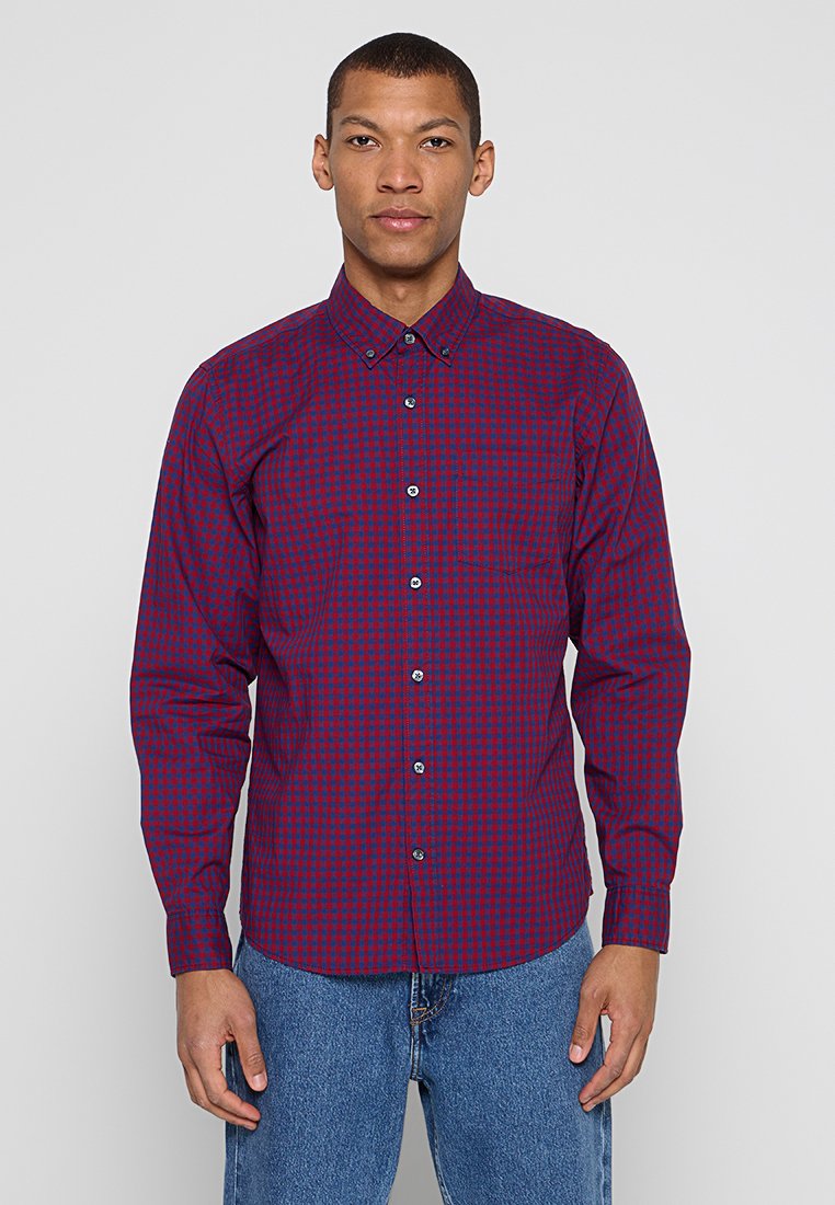Mann trägt ein rot-navy kariertes Hemd mit Knöpfen und blaue Jeans, steht vor einem einfachen hellen Hintergrund.
