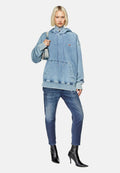 Junge Person mit kurzen blonden Haaren, die einen übergroßen blauen Denim-Hoodie, ausgebleichte enge Jeans und schwarze Absatzstiefel trägt und eine Umhängetasche hält.