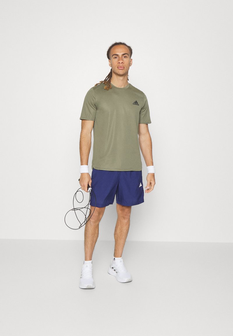 Olivegrünes sportliches T-Shirt, blaue Shorts, weiße Turnschuhe. Das Modell hält ein Springseil; mit Logo-Akzenten auf der Kleidung. Minimalistischer Hintergrund.