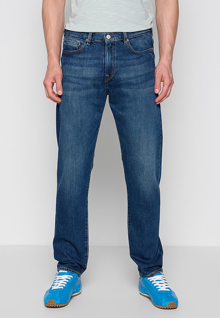 Dunkelblaue Jeans mit geradem Bein, fünf Taschen und kontrastierenden orangefarbenen Nähten, getragen mit blauen Sportschuhen.