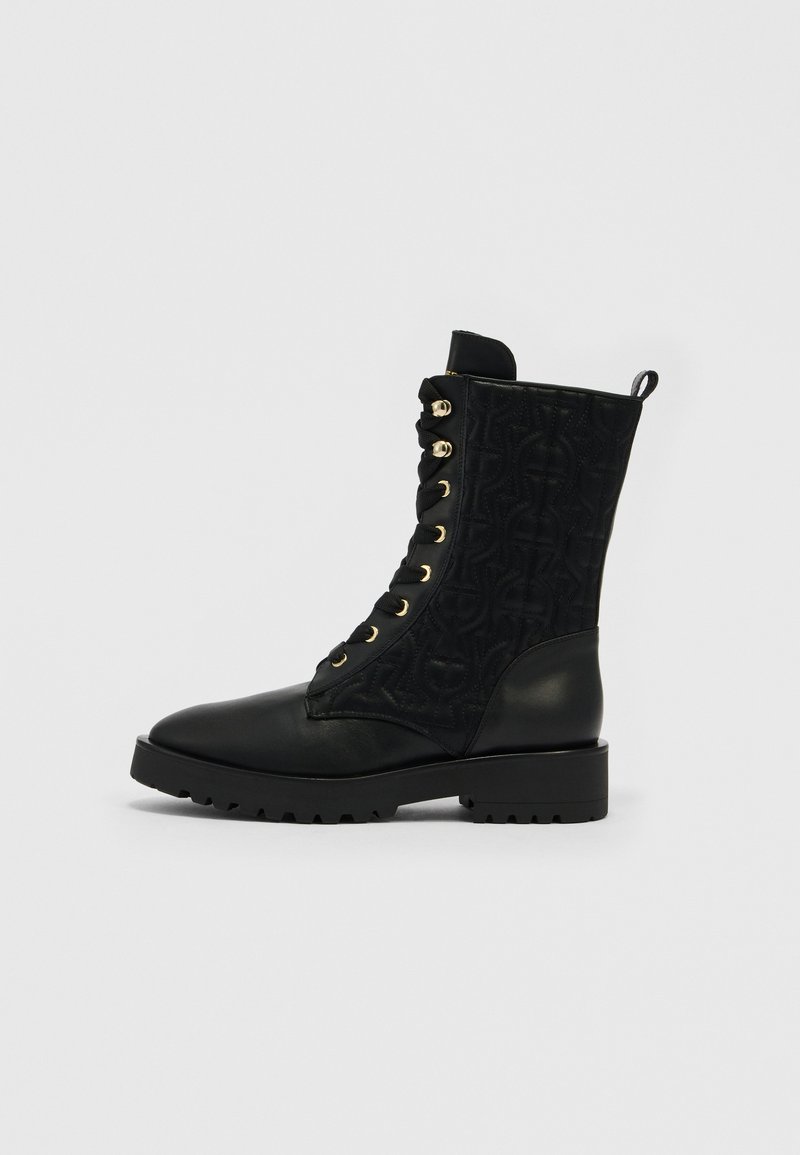 Schwarzer Kampfstiefel bis zur Mitte der Wade mit gesteppter Musterung aus Stoff, Lederzehen- und Fersenteil, goldfarbenen Ösen, schwarzen Schnürsenkeln und klobiger Profilsohle.