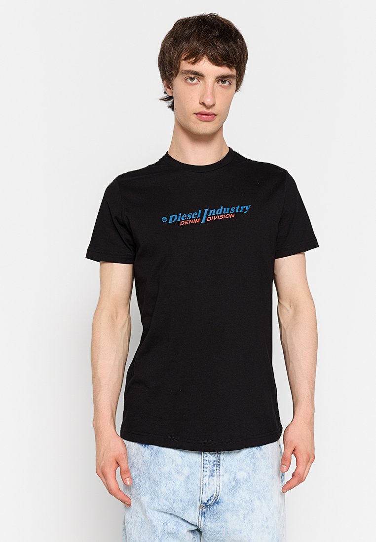 Junger Mann mit kurzen braunen Haaren, der ein schwarzes T-Shirt mit dem Diesel Industry-Logo und hellblauen Acid-Wash-Jeans trägt, steht vor einem weißen Hintergrund.
